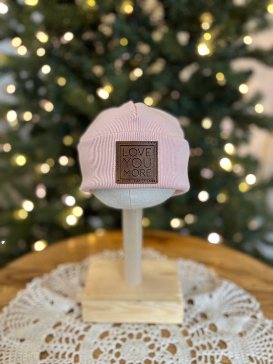 Pink cap with 'LOVE YOU MORE' text on a stand in front of a decorated Christmas tree.