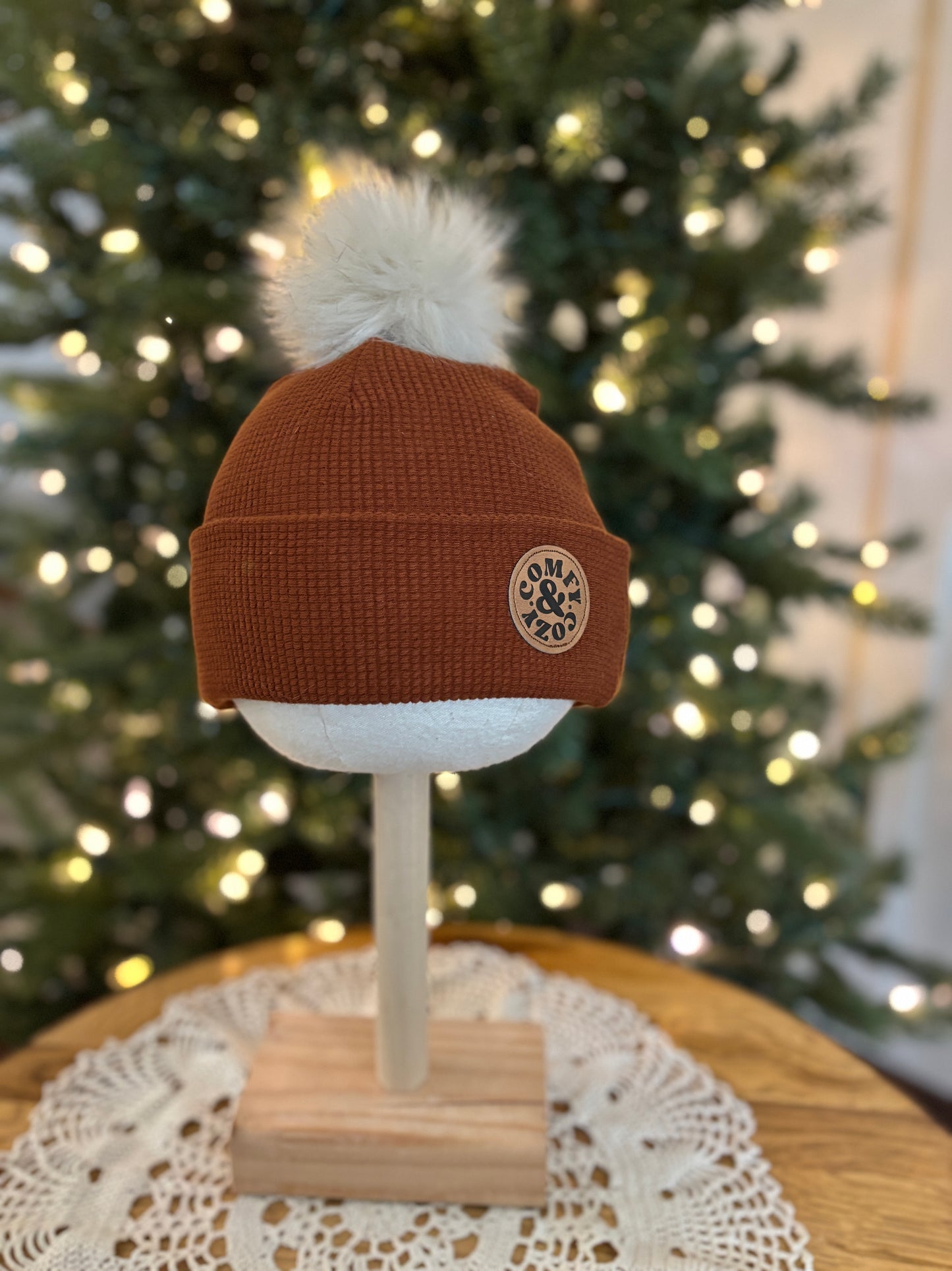 Brown beanie with a white pom-pom on a mannequin head in front of a Christmas tree.