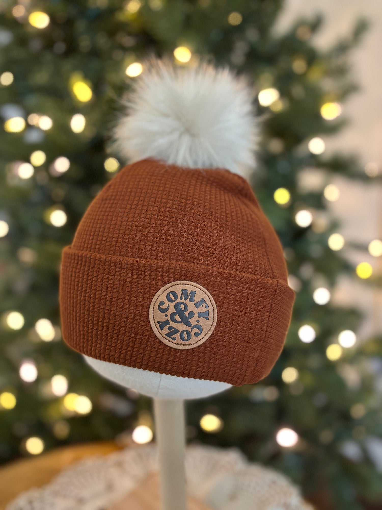 Brown beanie with white pom-pom and logo against a blurred Christmas tree background