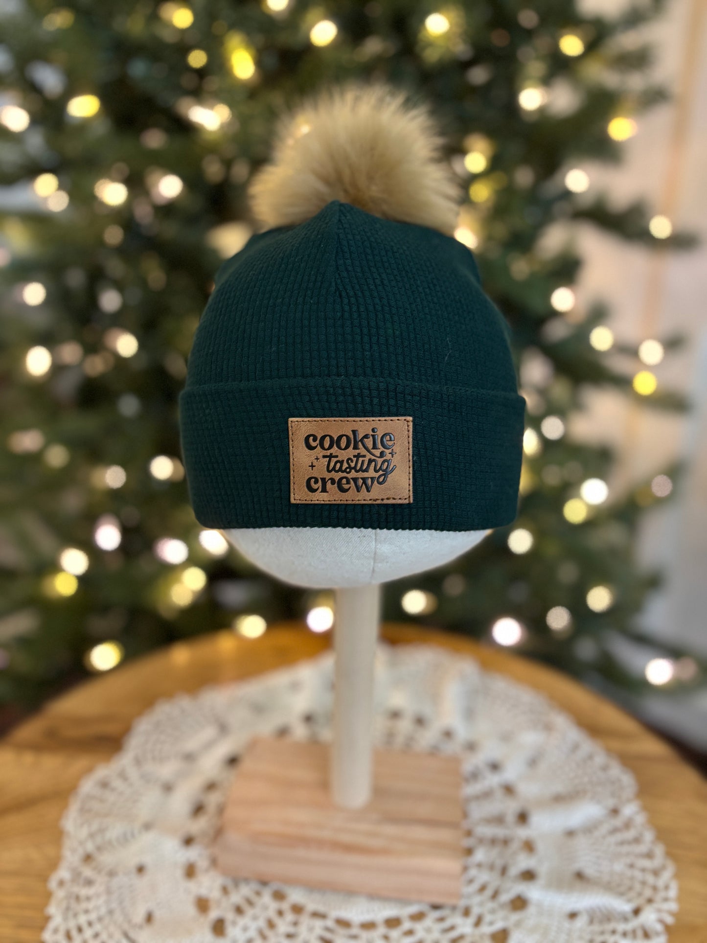 Dark green beanie with a pom-pom on a mannequin head in front of a decorated Christmas tree.