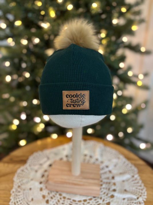 Dark green beanie with a pom-pom on a mannequin head in front of a decorated Christmas tree.