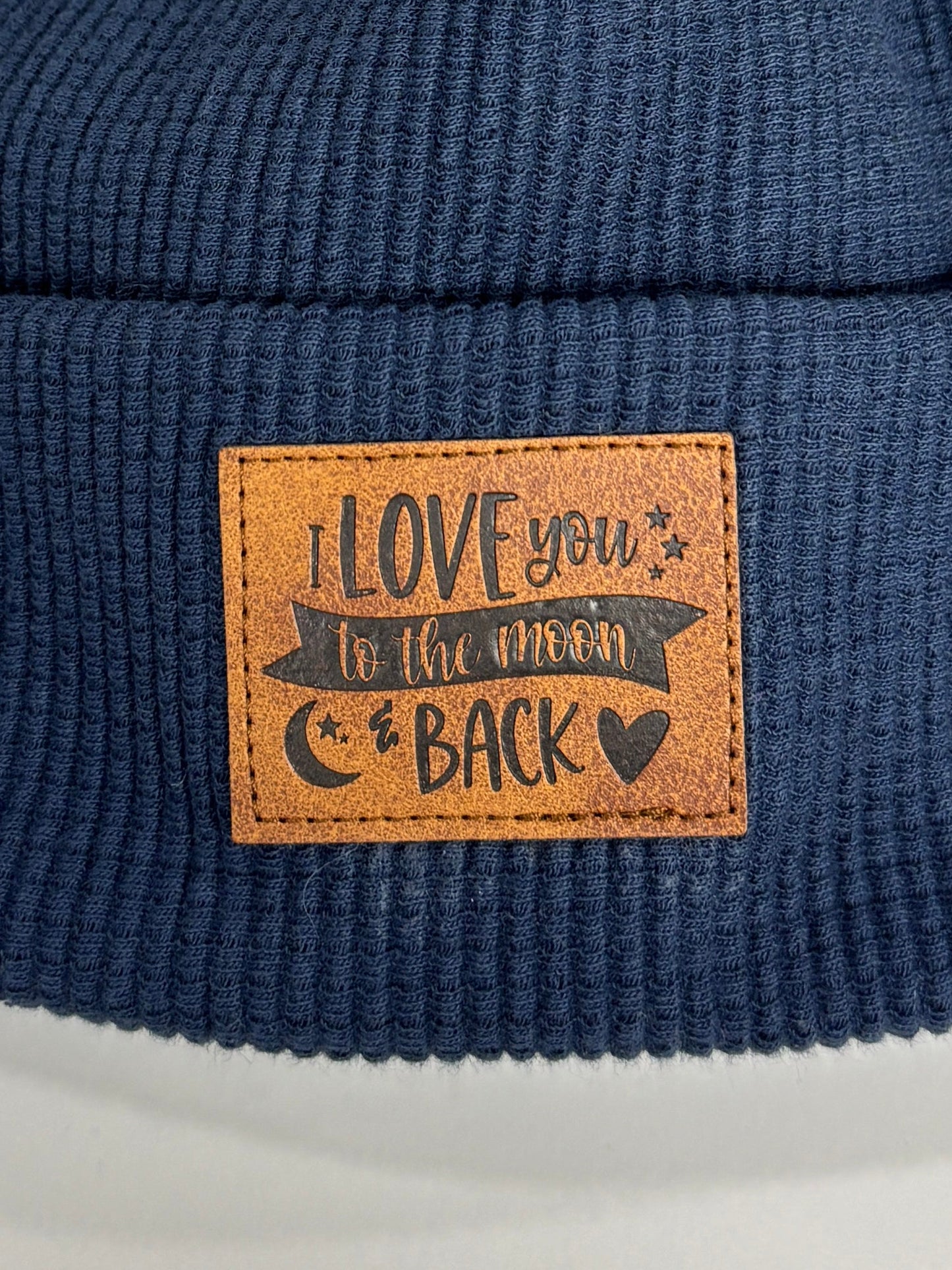 Navy blue beanie with a brown pom-pom and leather patch on a white background