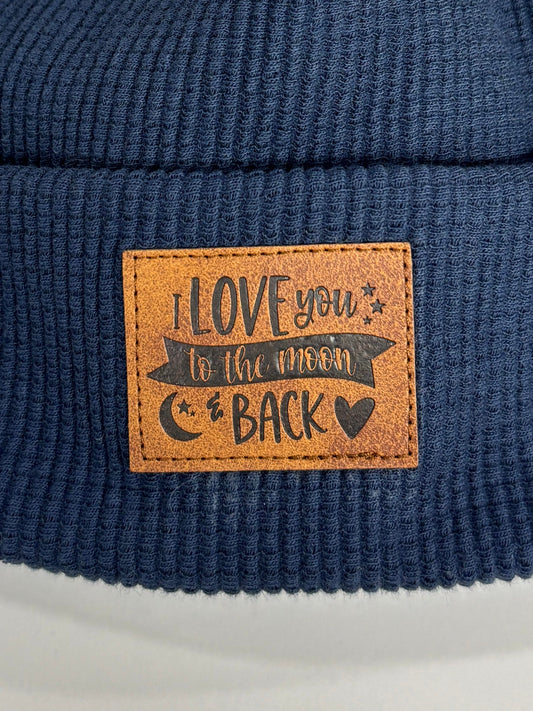 Navy blue beanie with a brown pom-pom and leather patch on a white background