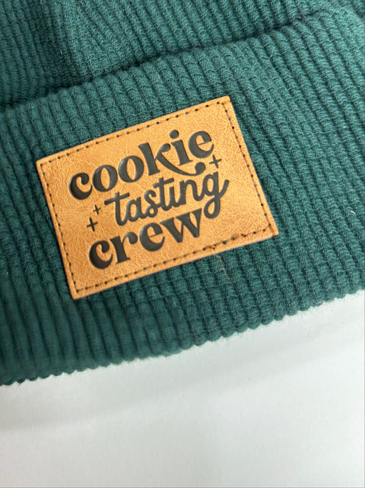 Green fabric with a brown leather label reading 'Cookie Tasting Crew' on a blurred background.